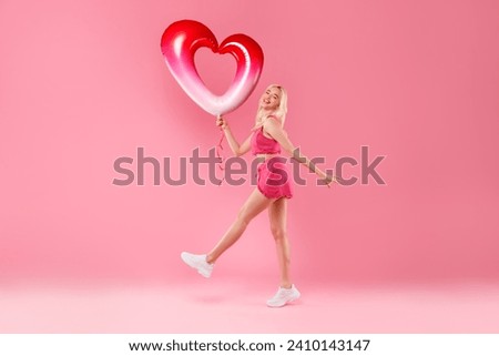 Blonde girl joyfully dancing, holding a red and white heart balloon, in pink fuzzy outfit with sneakers on a pink background, feeling elated, full length Royalty-Free Stock Photo #2410143147