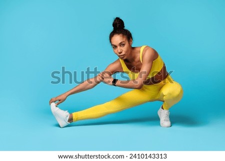 Athletic young black woman exercising during flexibility workout, stretching legs muscles, posing in extended leg squat position over blue studio backdrop, looking at camera. Sport, fitness Royalty-Free Stock Photo #2410143313