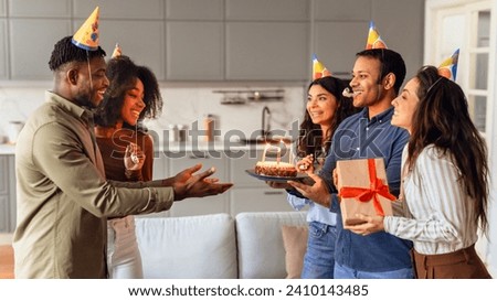 Joyful group of diverse friends giving birthday cake to a guy in festive hat, celebrating bday and making surprise party at modern apartment interior, presenting gifts in living room. Panorama