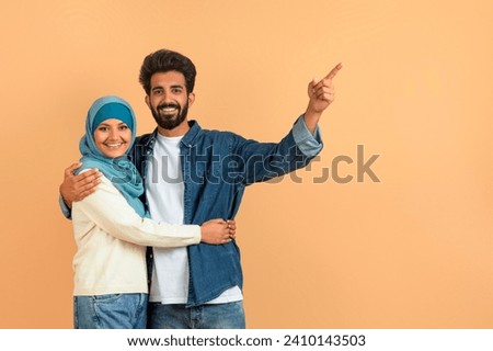 Arab muslim couple embracing and pointing aside at free space, smiling young islamic spouses sharing moment of discovery, demonstrating place for your ad, standing over soft beige studio background Royalty-Free Stock Photo #2410143503