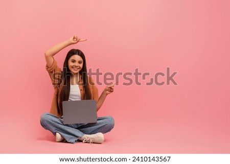 Cheerful Teen Girl Sitting On Floor With Laptop And Pointing Aside With Two Hands, Happy Female Teenager Demonstrating Copy Space For Advertisement Or Offer, Posing On Pink Studio Background
