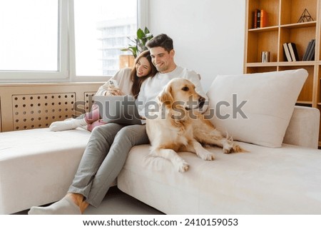 Portrait of happy young couple using laptop, man holding mobile phone, hugging dog, sitting together on comfortable sofa at home. Online shopping concept Royalty-Free Stock Photo #2410159053