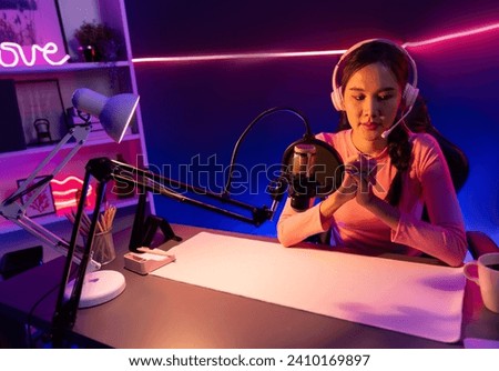 Smiling creative young beautiful Asian influencer talking in channel with positive podcast speech at night time, using laptop for presentation online at pink neon light modern studio. Stratagem.