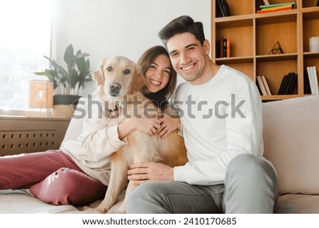Authentic portrait smiling couple, attractive happy man and woman hugging cute golden retriever dog  looking at camera sitting on comfortable sofa at cozy home. Love, wellbeing concept 