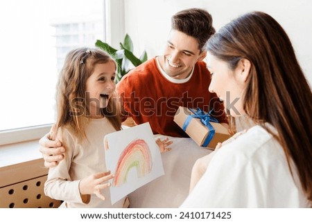 Portrait of happy young family. Smiling father, mother and little daughter holding drawing, giving gift, sitting in cozy room at home. Concept of holiday, birthday, mother's day