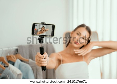 Young woman making fashion video content for social media. Blogger smiles to camera using selfie stick and light ring while making persuasive online clothing sell to audience or followers. Unveiling Royalty-Free Stock Photo #2410176671