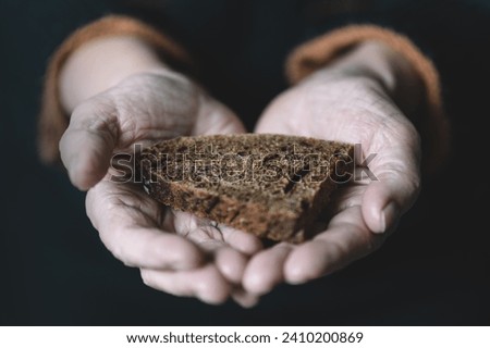an old retired woman holds in her hand a piece of black rye bread on a dark background. The concept of hunger and poverty.