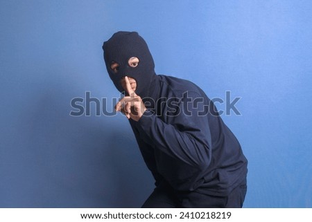 Thief in black balaclava holding fingers on lips, making hush sign isolated on blue background Royalty-Free Stock Photo #2410218219