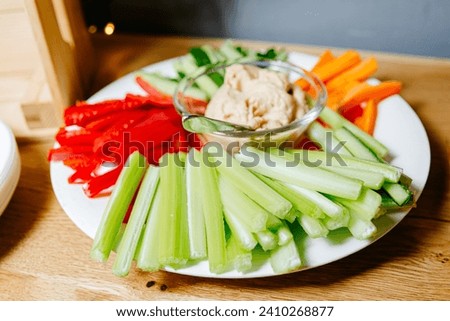 Colorful vegetable snack platter with fresh celery sticks, carrot sticks, red bell pepper, and creamy hummus dip in a glass bowl. Royalty-Free Stock Photo #2410268877