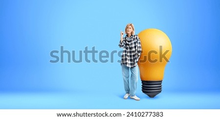 Woman finger pointing up, full length standing near large lightbulb on empty blue background. Concept of inspiration, start up, creativity, knowledge and business idea