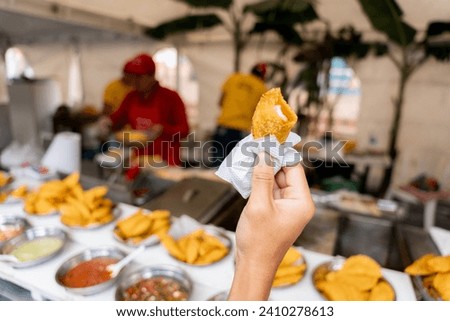 A close-up view of a hand holding a traditional Latin empanada in front of a street food stall Royalty-Free Stock Photo #2410278613