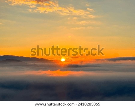 This image shows a sunrise over a sea of clouds. The sun is shining brightly and the sky is a beautiful orange. The clouds are fluffy and white and they stretch out as far as the eye can see