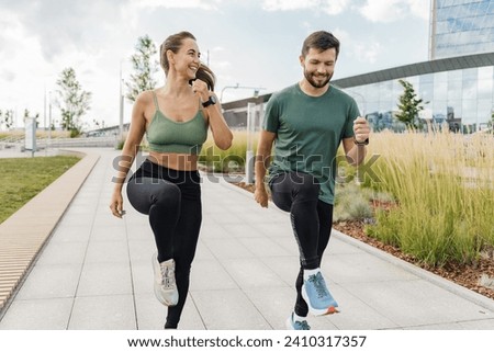 Energetic couple high-knee jogging in city park, tall glass buildings in the distance. Royalty-Free Stock Photo #2410317357