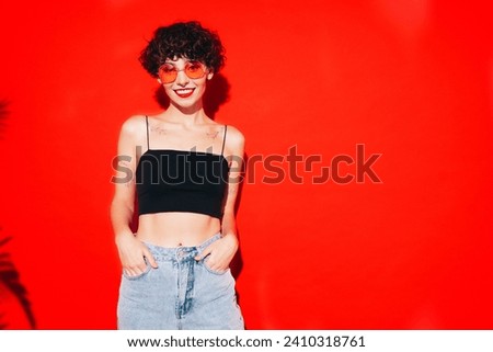 Young beautiful smiling female in trendy summer black tank top. Carefree woman posing near red wall in studio with curly hairstyle. Positive model having fun. Cheerful and happy. In sunglasses