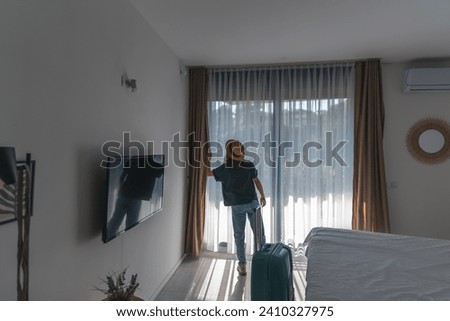 A young woman in a hat with a suitcase checks into a hotel room. Booking and Accommodation travel  Royalty-Free Stock Photo #2410327975