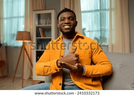 Portrait of smiling happy African American man speaking with sign language, showing finger symbols, wearing casual clothes, looking at camera while sitting at home. Communication concept