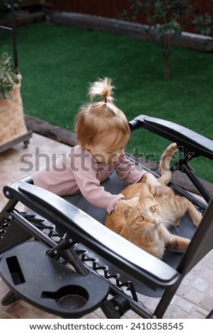 Little red-haired charming girl in the garden tries to pet a cat sitting on a garden chair