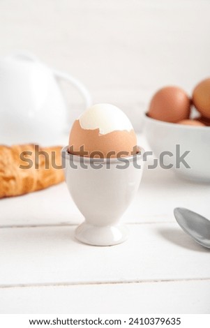 Holder with boiled chicken egg, teapot and croissant on white wooden background