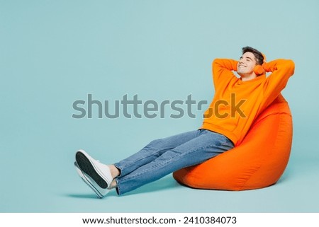 Full body young man he wearing orange hoody casual clothes sit in bag chair listen to music in headphones isolated on plain pastel light blue cyan color background studio portrait. Lifestyle concept