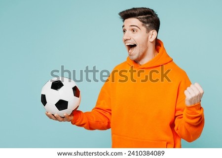 Young excited man fan he wears orange hoody casual clothes do winner gesture cheer up support football sport team hold in hand soccer ball watch tv live stream isolated on plain blue color background