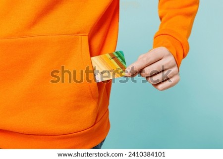 Close up cropped photo of young man he wears orange casual clothes hold in hand put into pocket mock up of credit bank card isolated on plain blue cyan color wall background studio. Lifestyle concept