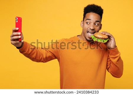 Young man wear orange sweatshirt casual clothes hold eat bite burger do selfie shot on mobile cell phone isolated on plain yellow background Proper nutrition healthy fast food unhealthy choice concept