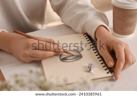 Woman drawing cup of drink with graphite pencil in sketchbook at table, closeup