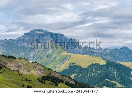 Panoramic view of mountain ridges and slopes with green pastures in summer and cloudy sky in the background. A view to Gsür 2708 m., a mountain in the Bernese Alps, Adelboden, Switzerland. Royalty-Free Stock Photo #2410447177