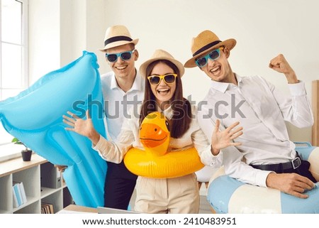 Team of happy corporate employees going on summer holiday together. Group of three cheerful, joyful young people, ready for cool vacation, having fun with inflatable toys and mattress in the office