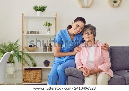 A beautiful, young, happy, smiling nurse kindly embraces her patient, an elderly, well-groomed, pretty gray-haired lady with glasses, sitting down on the edge of the sofa for photo posing for memory. Royalty-Free Stock Photo #2410496323