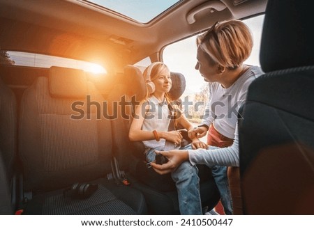 Inside the car photo of a mother fastening with safety auto belt her little daughter girl sitting in child seat. Girl listening music using headphones. Family values, traveling and technology concept Royalty-Free Stock Photo #2410500447