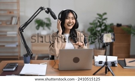 Woman host working on laptop with microphone and recording audio podcast on smartphone in studio.