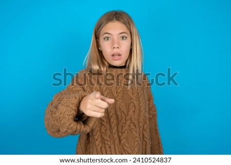 Shocked beautiful caucasian teen girl wearing brown sweater points at you with stunned expression