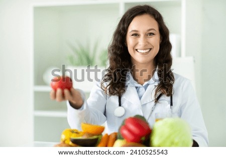 Young european woman dietitian holding apple, sitting at desk with fruits and vegetables, smiling at camera. Lady nutritionist recommending weight loss clinic, copy space Royalty-Free Stock Photo #2410525543