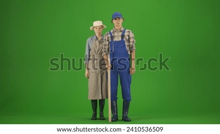 Portrait of farmer in working clothing on chroma key green screen. Gardener man and woman with pitchfork posing holding hands.
