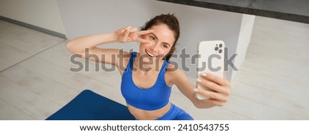 Portrait of young styling fitness girl doing workout from home, taking selfie and video for social media, gym instructor records her training session indoors.