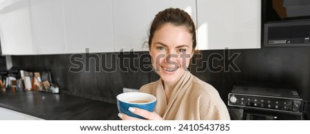 Good-looking young woman with cup of coffee, posing in the kitchen, smiling at camera, enjoys her morning at home, wearing cosy bathrobe.