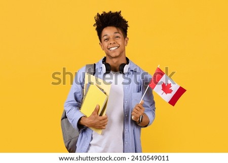 Cheerful black male student proudly holding Canadian flag, wearing headphones, ready for study abroad, against yellow background, symbolizing diversity and foreign languages learning