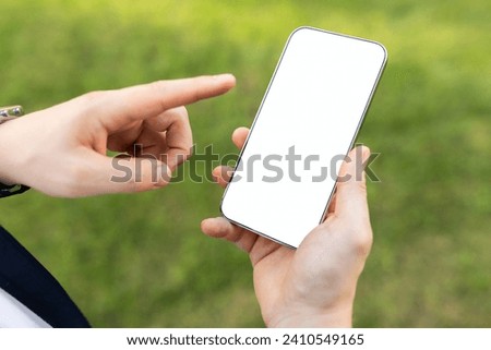 Close-up of a person hands holding smartphone with blank white screen, pointing with finger, with green grass background, close up, cropped. App, website, blog recommendation