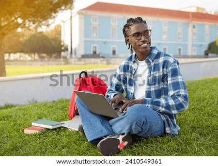 Black young man student working on laptop computer, websurfing for university lectures in academic pursuit, seated outdoors on backdrop of college building, studying at campus park Royalty-Free Stock Photo #2410549361