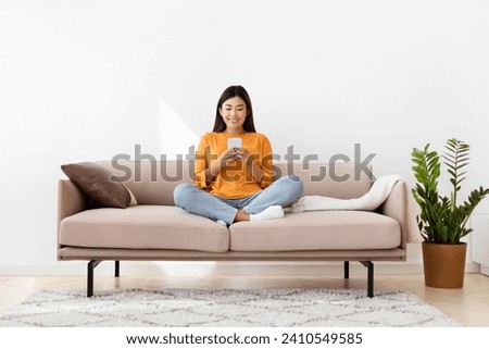 Smiling millennial asian lady holding smartphone, sitting on couch in cozy living room, home interior. Filipino woman reading online book or blog on cell phone, copy space, full length Royalty-Free Stock Photo #2410549585