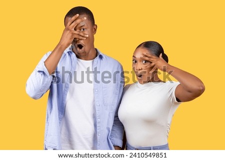 Black man and woman display humorous shock, covering their eyes with their hands while peeking through, creatin playful and surprised atmosphere on yellow background