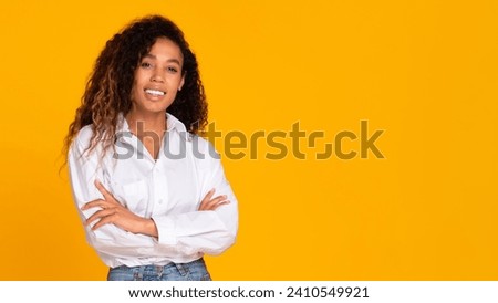 Black young lady with curly hair smiling and crossing hands, stands confidently over yellow studio background, expressing confidence and beauty. Panorama, free space for text