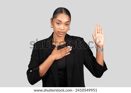 Sincere African American businesswoman making a pledge or swearing an oath with her right hand raised and left hand on her chest, in a professional black suit on grey background, studio Royalty-Free Stock Photo #2410549951