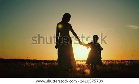 Silhouettes of mamma holding hand of daughter and talking about childhood while walking with child in sunlit field. Silhouettes of mamma with daughter sharing secrets at setting sun in park