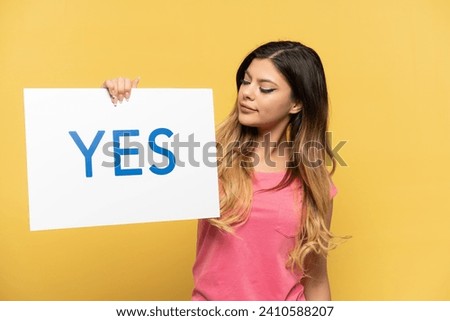 Young Russian girl isolated on yellow background holding a placard with text YES