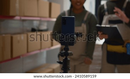 Warehouse logistics coordinator and colleague using smartphone placed on tripod to make training video for interns. Employees film themselves in fulfillment center showing trainees how to seal boxes