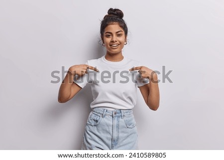 Horizontal shot of pretty dark haired woman with hair bun points index fingers on blank space of t shirt suggests to place your logo here isolated over white background. Clothes template concept Royalty-Free Stock Photo #2410589805