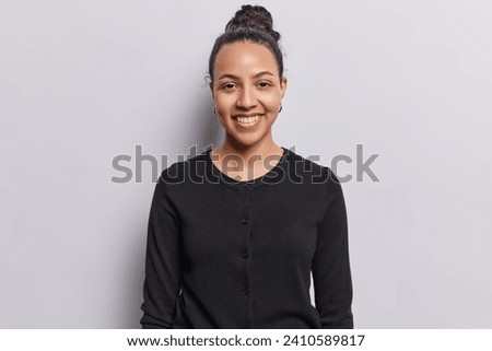 Waist up shot of cheerful Latin woman smiles gently looks directly at camera dressed in casual black jumper isolated over white background. Beautiful young smiling female model being in good mood Royalty-Free Stock Photo #2410589817