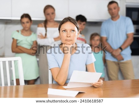 Frustrated woman sits in kitchen and reads unpleasant news in letter. Rent increase, penalty for late payment of goods, business problems. Blurred family husband and children in background Royalty-Free Stock Photo #2410618587